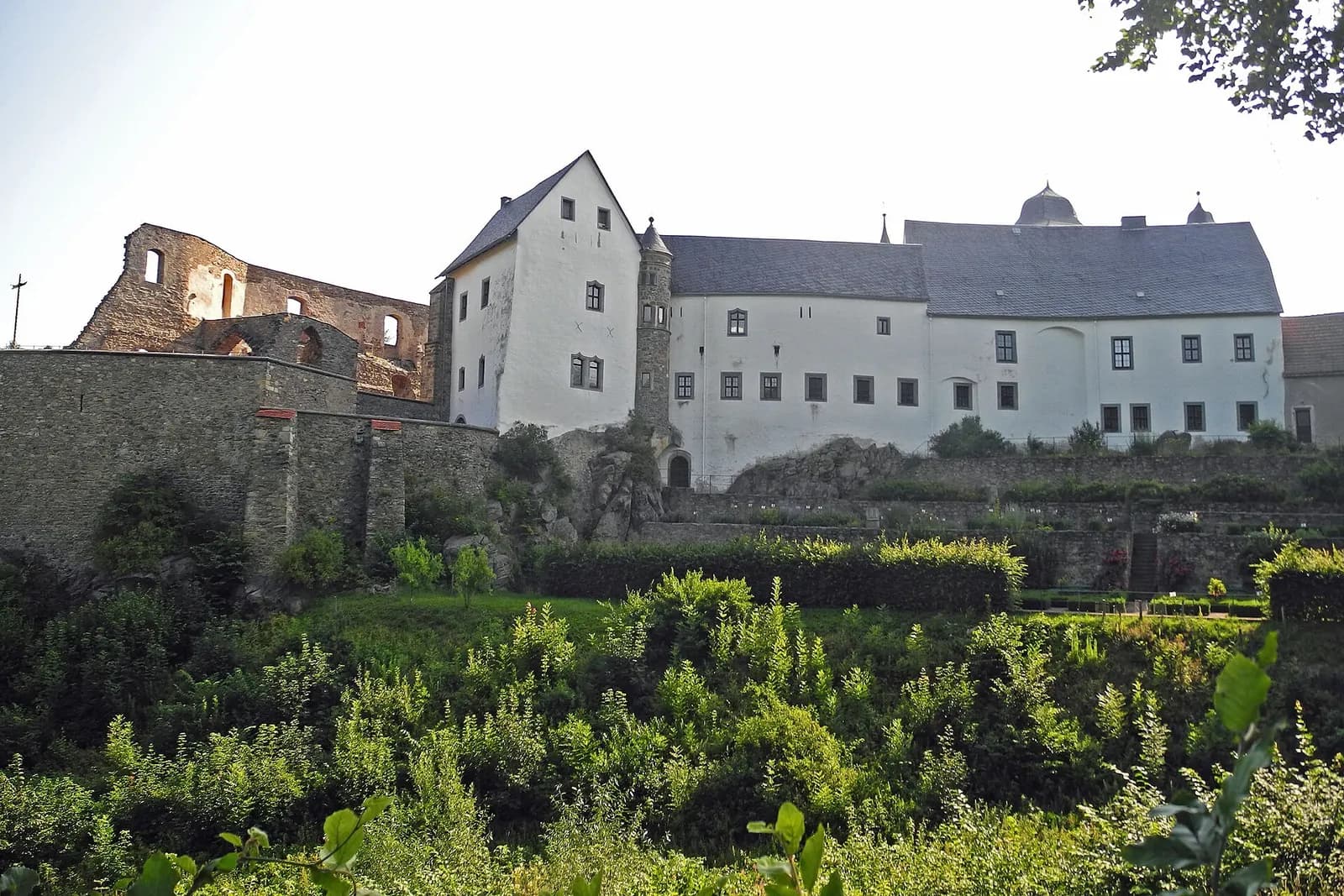 Weißes Schloss Lauenstein mit Türmchen und Burgruine auf einem Felsen, davor grüner Hang