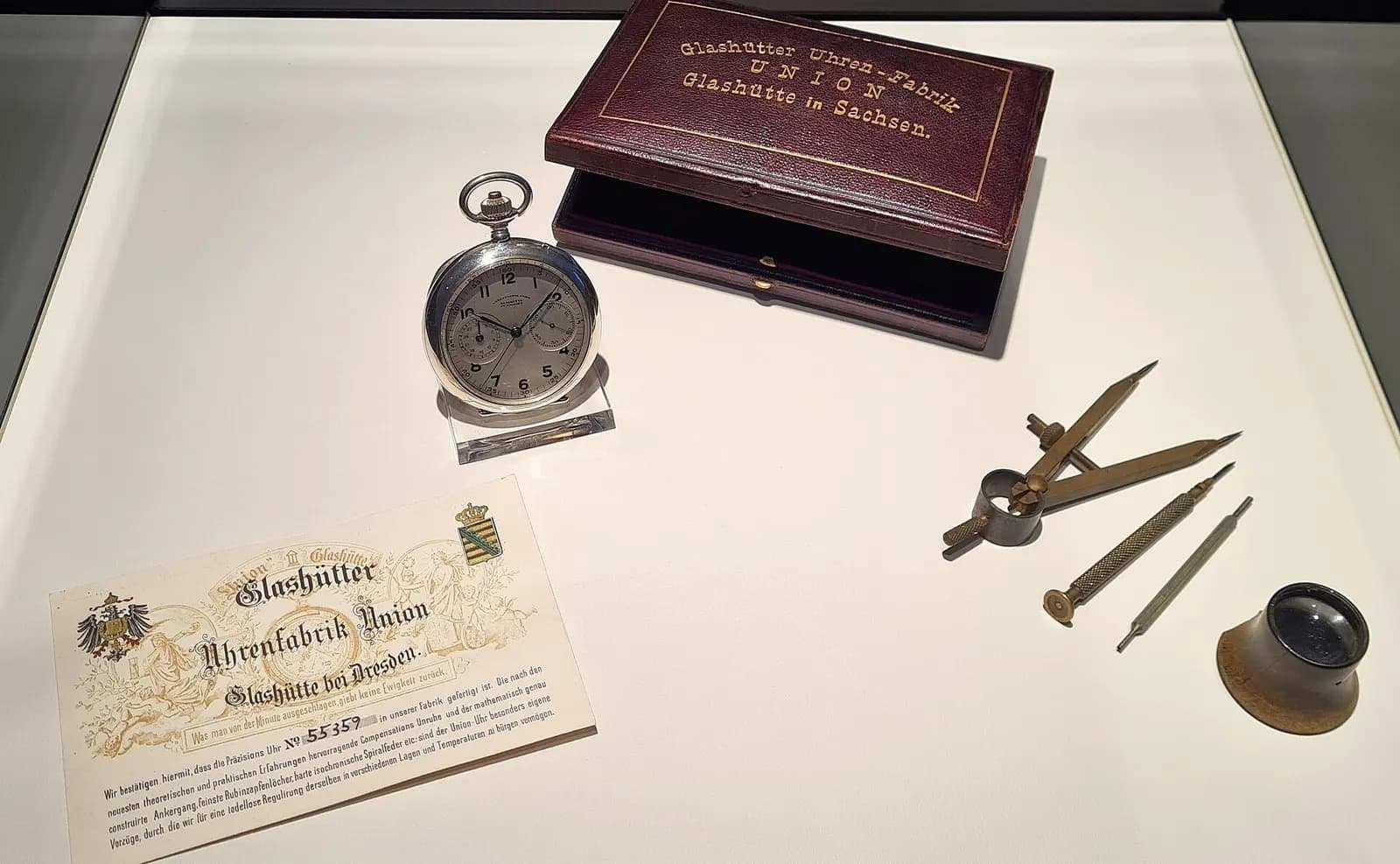 Taschenuhr der Glashütter Uhrenfabrik Union in einer Vitrine, daneben Etui, Zertifikat und Uhrmacherwerkzeug