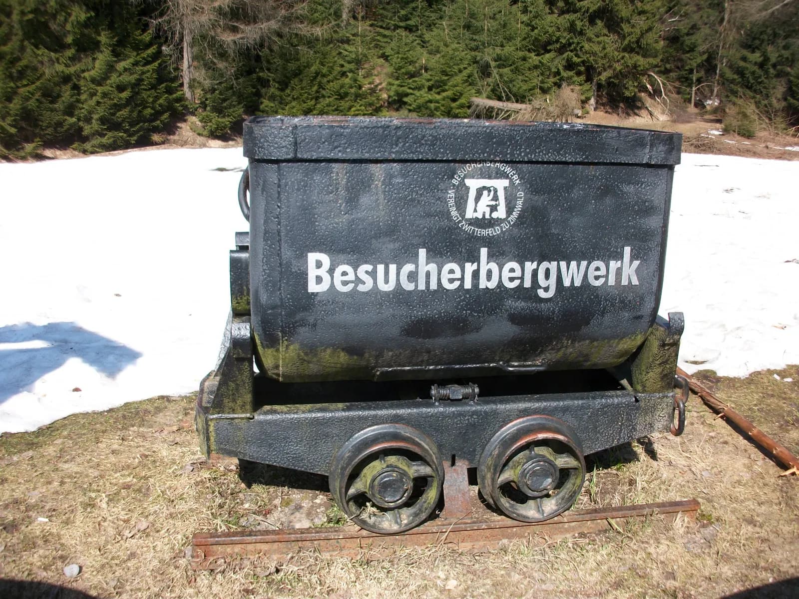 Schwarzer Förderwagen mit der Aufschrift Besucherbergwerk auf Schienen, daneben Schneereste und Fichtenwald