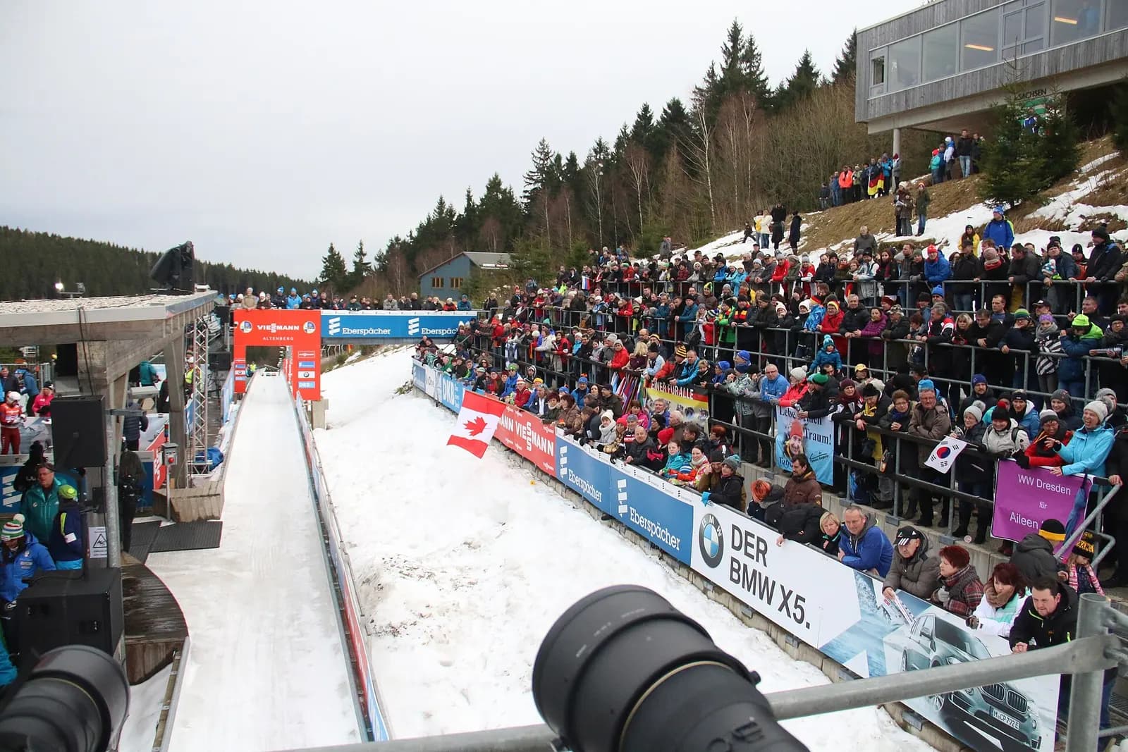 Zuschauer drängen sich hinter der Bande am Zielbereich des Eiskanals, daneben eine Kameraposition