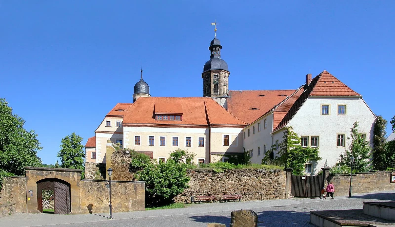 Schloss Dippoldiswalde mit hellem Hauptbau, rotem Dach und Kirchturm, davor alte Mauer mit Torbogen