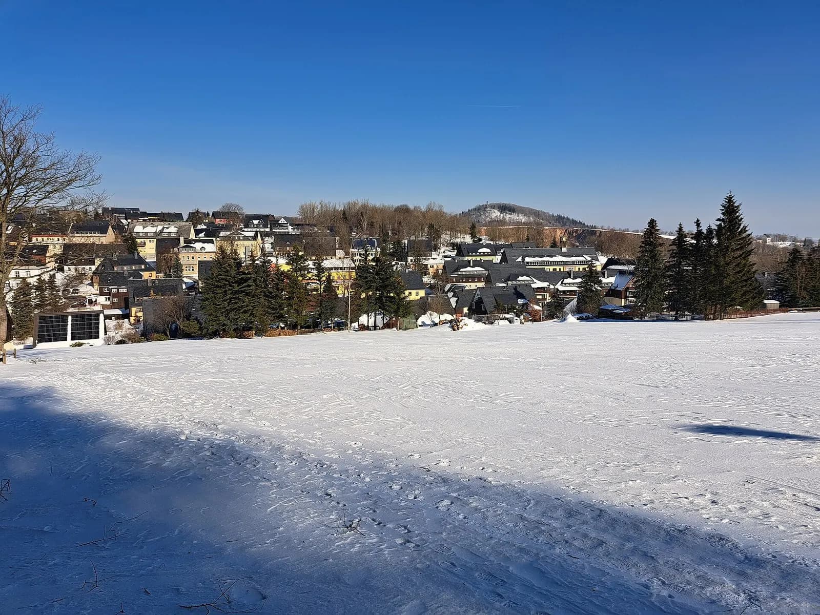 Verschneite Wiese am Ortsrand von Altenberg unter blauem Himmel, im Hintergrund die Häuser des Ortes
