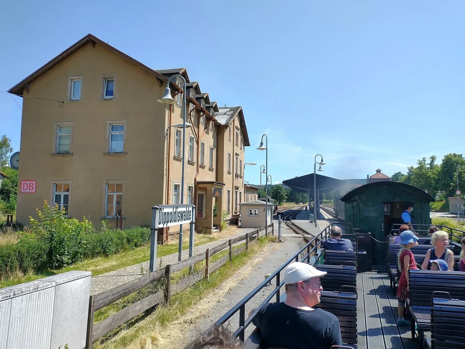 Offener Aussichtswagen der Weißeritztalbahn mit Fahrgästen im Bahnhof Dippoldiswalde, dahinter das gelbe Empfangsgebäude
